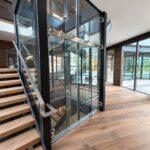 Custom glass elevator with black steel shaft beside an oak staircase in a modern luxury residence