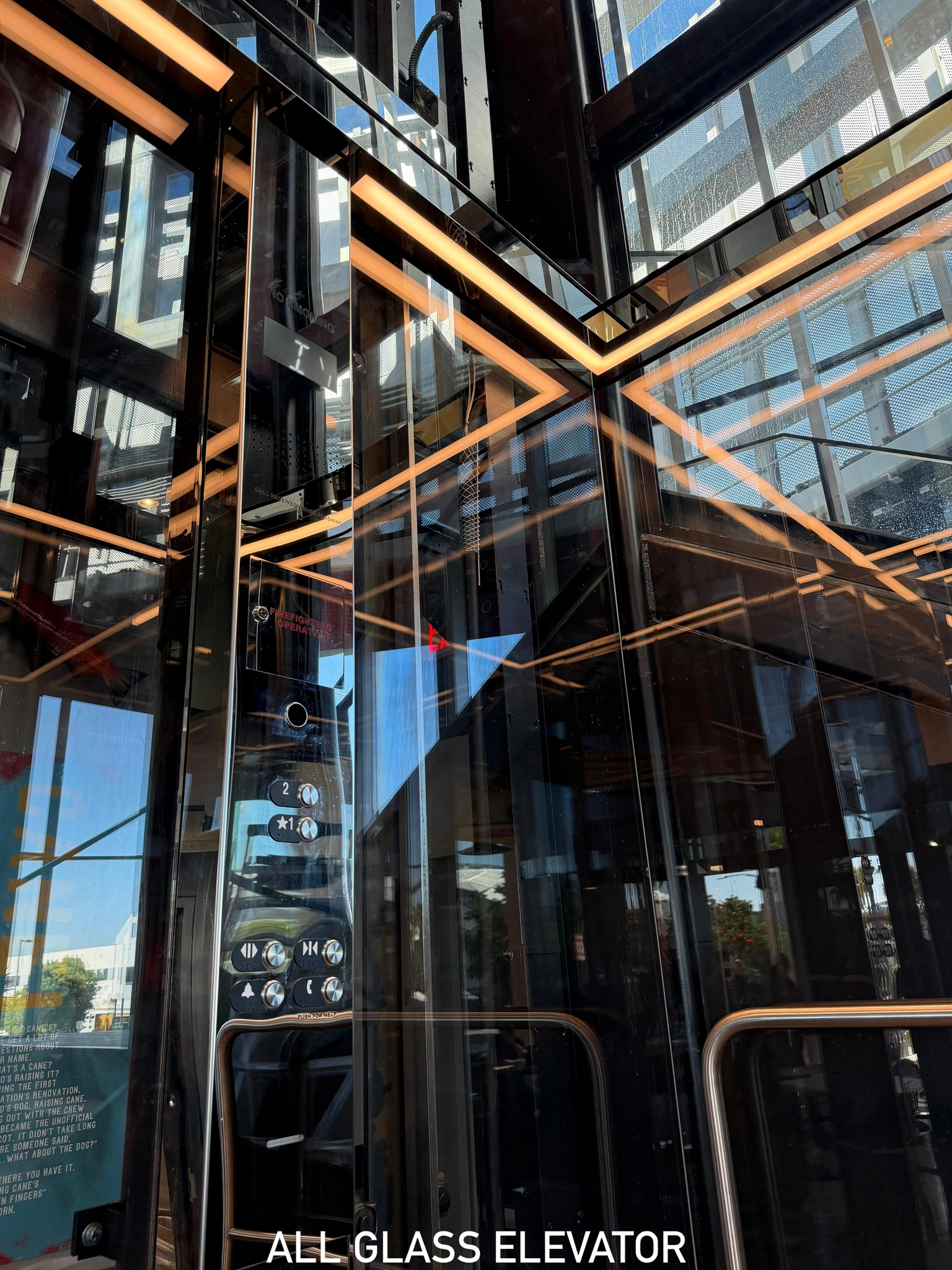 Glass elevator interior with transparent walls, polished metal fittings and warm linear lighting reflected in the glazing.