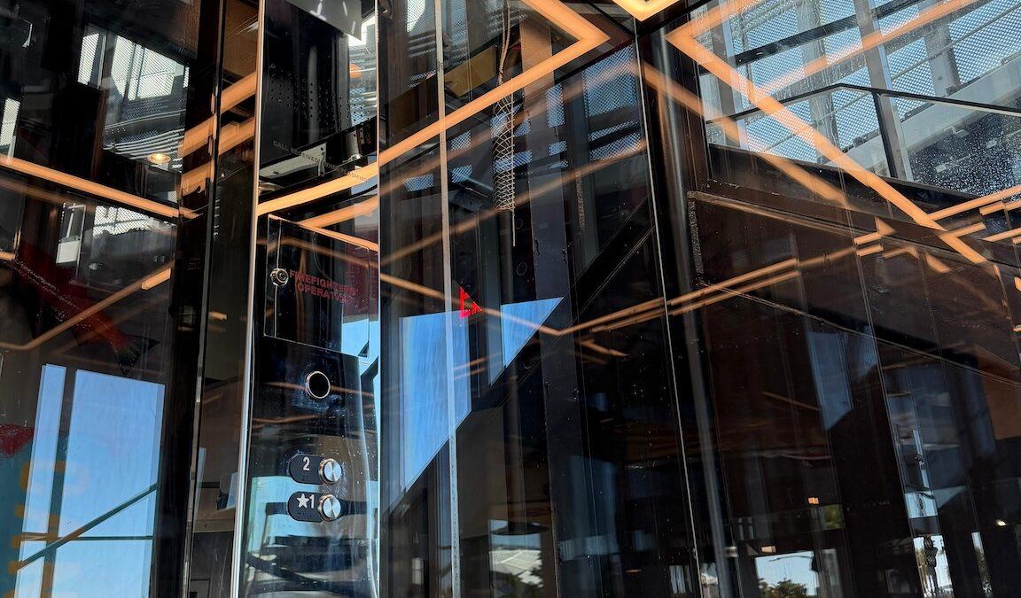 Glass elevator interior at Raising Cane's Inglewood flagship, with transparent walls and warm linear ceiling lighting.