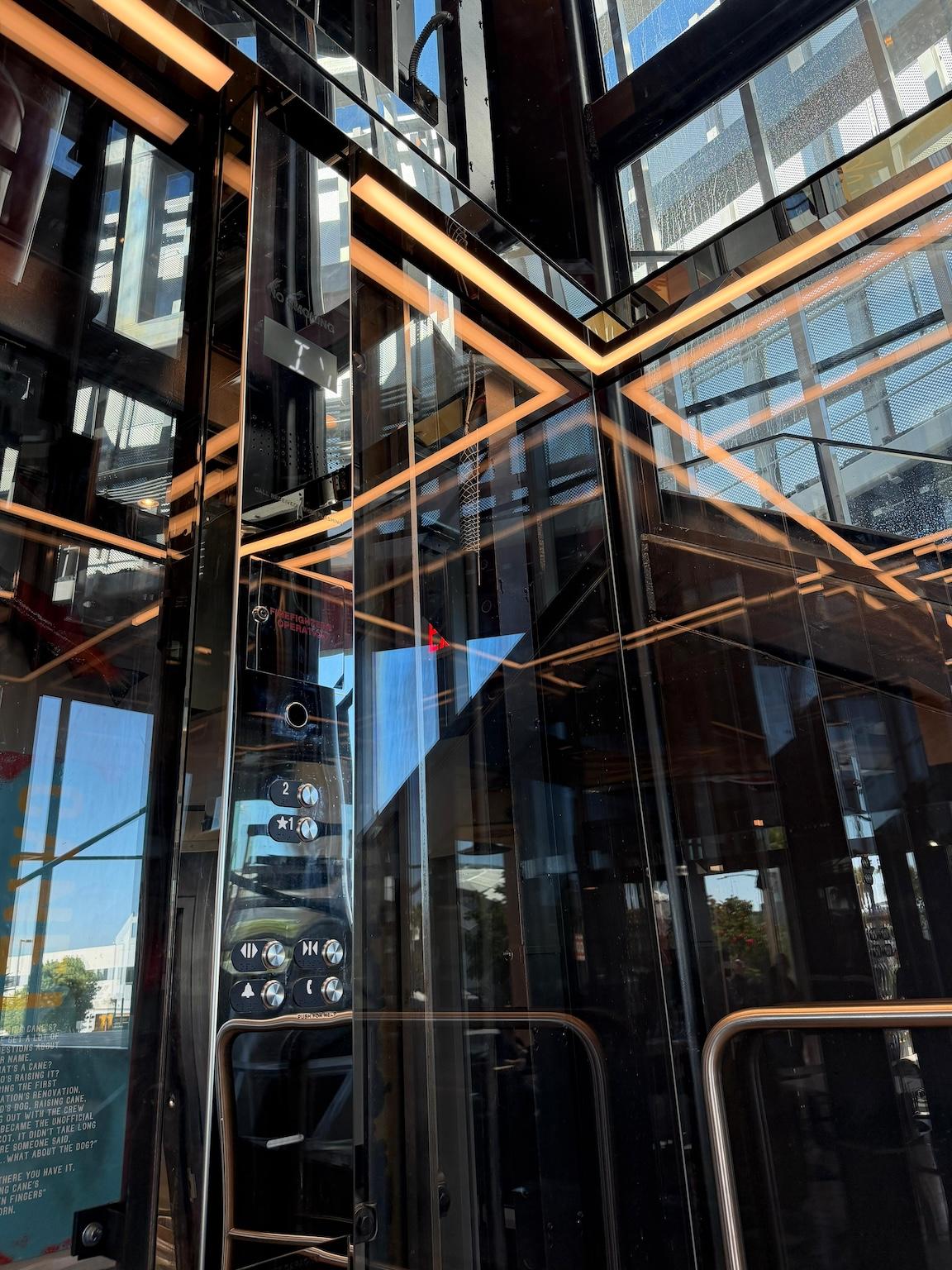 Glass elevator interior at Raising Cane's Inglewood flagship, with transparent walls and warm linear ceiling lighting.